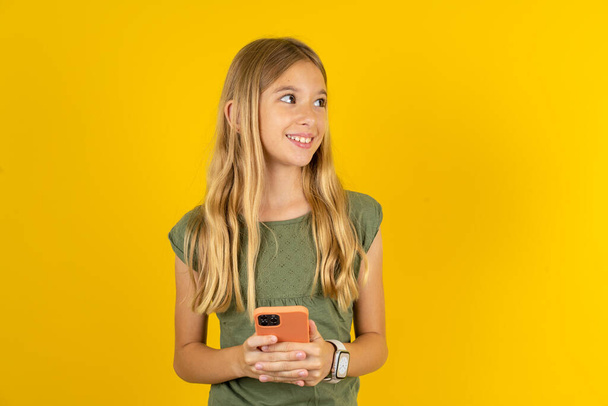 blond klein meisje dragen kaki blouse over gele achtergrond houden telefoon handen lezen goed jeugd nieuws kijken leeg ruimte advertentie - Foto, afbeelding