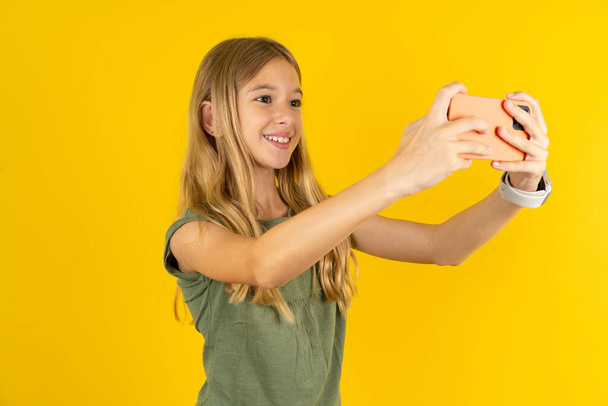 blond little girl wearing khaki blouse over yellow background taking a selfie to post it on social media or having a video call with friends. - Photo, Image