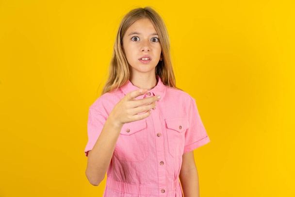 Шокированная девушка в розовой куртке на желтом фоне указывает на тебя с ошеломленным выражением лица - Фото, изображение