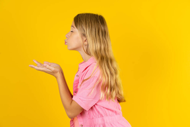 Profile side view, portrait of attractive girl wearing pink jacket over yellow background sending air kiss - Photo, Image
