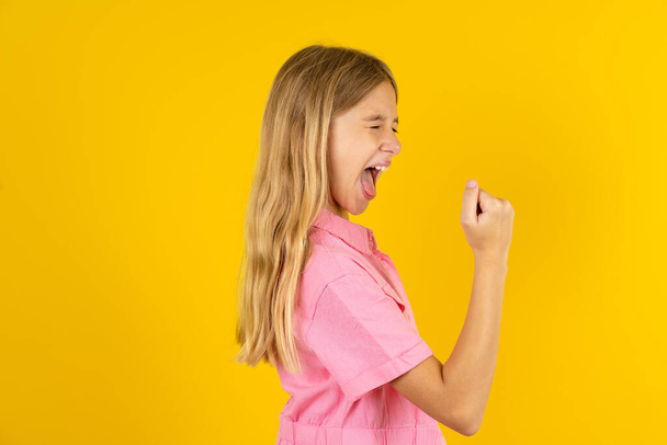 Porträt von lustigen Mädchen in rosa Jacke über gelbem Hintergrund schreien yeah erhobene Fäuste Hände feiern Sieg Spiel Wettbewerb - Foto, Bild