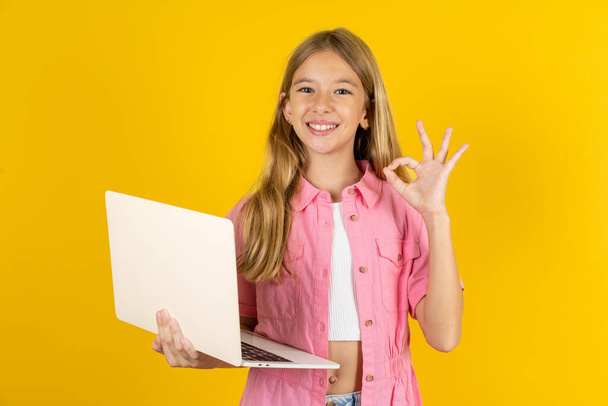 Chica positiva con chaqueta rosa sobre fondo amarillo mantenga los dedos inalámbricos netbook mano muestran símbolo okey - Foto, imagen