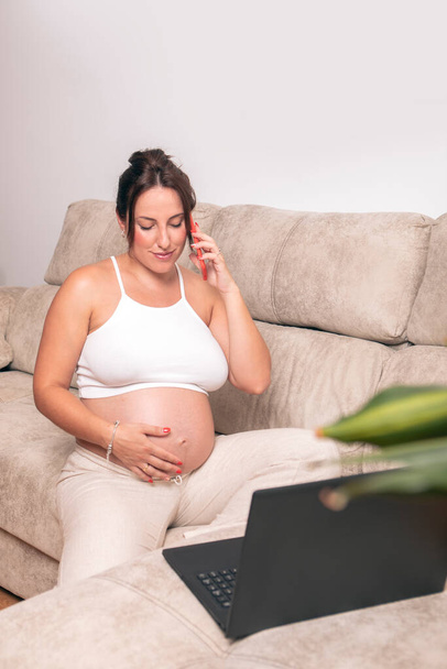 Une jeune femme enceinte travaille avec un ordinateur portable sur son canapé à la maison, tout en parlant au téléphone et en touchant son ventre. Photo de haute qualité - Photo, image