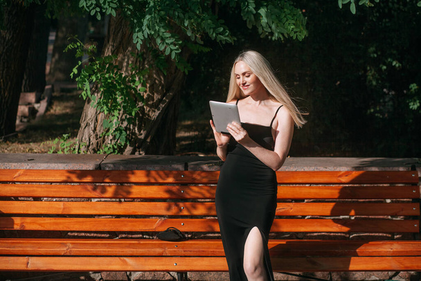 A positive pretty woman of the age with long blond hair, in a black long dress works remotely on a tablet, stands near a wooden bench in a city park, on a bright sunny day. - Photo, Image