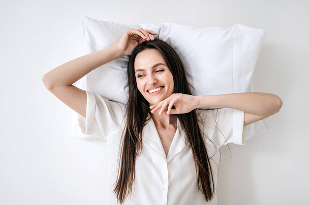 acima vista sobre a jovem mulher no pijama descansando sobre travesseiro confortável. sorridente fêmea acordar e sorrir largo, desfrutando de manhã isolado em fundo espaço cópia branca, estúdio tiro - Foto, Imagem