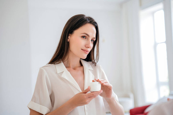 Portrait of woman in casual clothes holding jar with moisturizer cream, standing in white bathroom interior. Professional face lifting cosmetics for home spa and skincare routine. Advertising concept - Foto, imagen