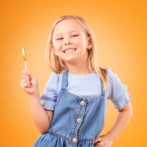 Kind, Lutscher oder Bonbonporträt im Atelier für Naschkatzen, Farbspirale oder Zucker für Energie. Gesicht des glücklichen Mädchens Kind auf orangefarbenem Hintergrund für Snack, aufgeregtes Lächeln und Dessert oder ungesundes Essen. - Foto, Bild