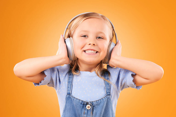 Ακουστικά, πορτρέτο ή παιδική μουσική streaming για να χαλαρώσετε με την ελευθερία στο στούντιο σε πορτοκαλί φόντο. Πρόσωπο, χαμόγελο ή ευτυχισμένη κοπέλα ακούγοντας ένα διασκεδαστικό τραγούδι ραδιόφωνο, ήχο ή ήχο σε μια online συνδρομή. - Φωτογραφία, εικόνα