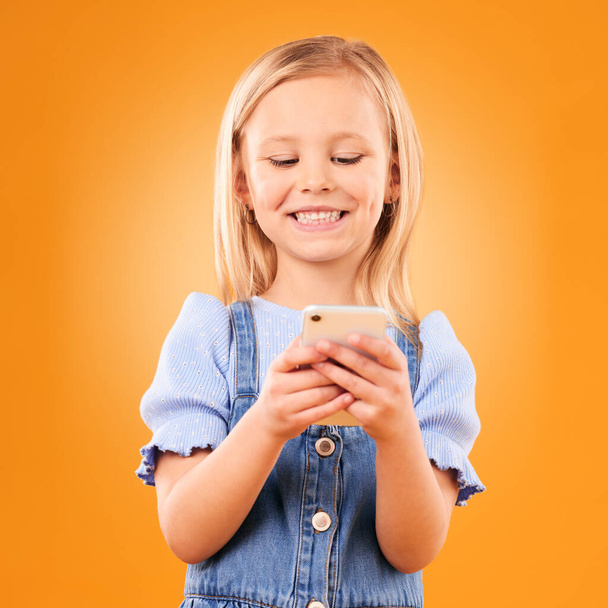Child, smile and typing with phone in studio for social media, play mobile games and download app on orange background. Happy girl kid, smartphone and reading notification, watch multimedia and chat. - Photo, Image