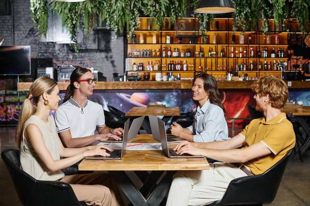 hardwerkende vrolijke team met behulp van laptops en glimlachen naar elkaar met wazig decor, coworking - Foto, afbeelding