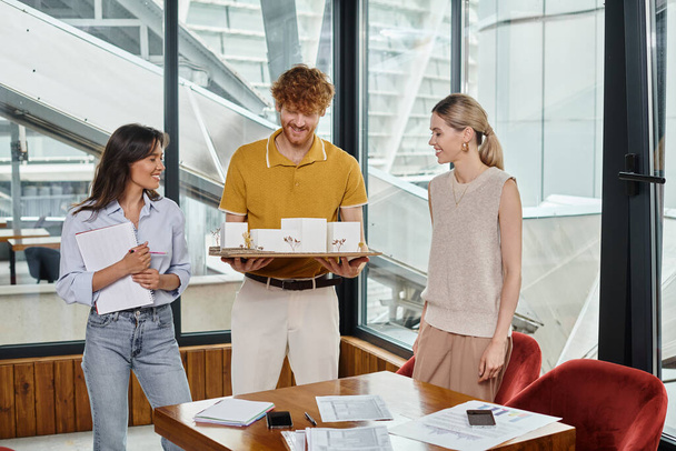 kolme iloinen nuori tiimin jäsentä hymyilee ja tarkastellaan mittakaavassa malli rakennuksen, suunnittelu toimisto - Valokuva, kuva