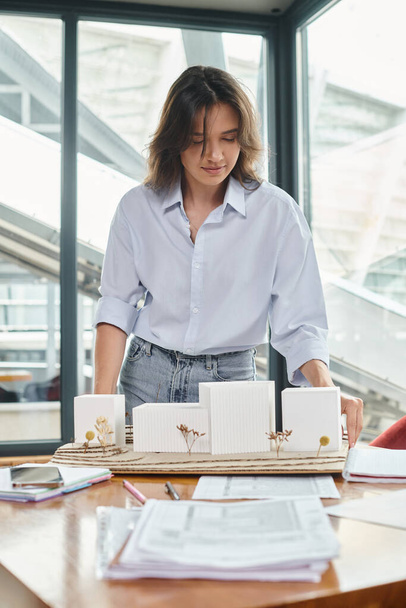 jong brunette vrouw werken aan haar documenten en schaal model met glas op de achtergrond, design bureau - Foto, afbeelding