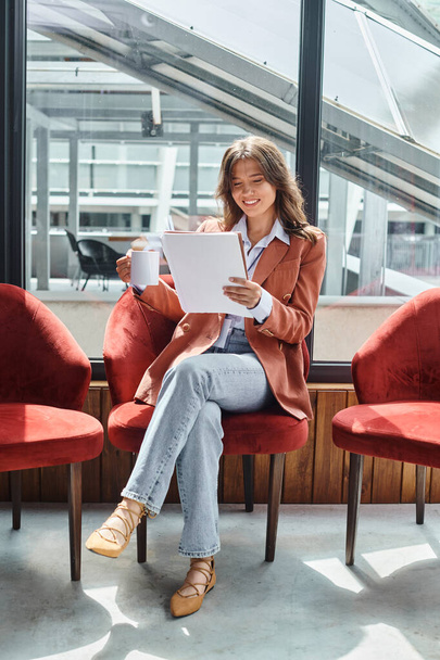 sorridente giovane donna in giacca marrone controllare la sua documentazione mentre prende un po 'di tè, coworking - Foto, immagini