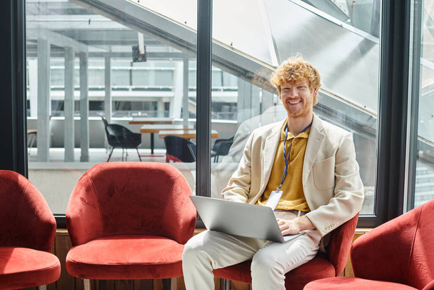 homme aux cheveux rouges joyeux avec ordinateur portable sur ses tours assis sur une chaise avec fond de verre, coworking - Photo, image