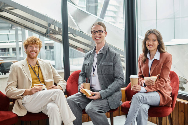 drei gut gelaunte Kollegen blicken in die Kamera und genießen Sandwiches und Kaffee, Coworking-Konzept - Foto, Bild