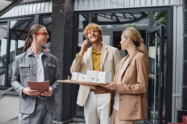 трое молодых коллег держат бумаги и масштабную модель разговора на мобильном телефоне, конструкторском бюро - Фото, изображение