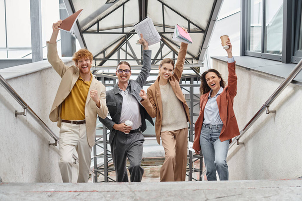 happy cheering teammates posing on staircase with hands with paperwork raised in air, coworking - Photo, Image