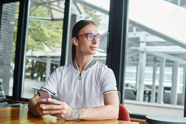 skupiony młodzieniec w okularach z telefonem i tatuażem odwracający wzrok szklanym tłem, współpracownik - Zdjęcie, obraz
