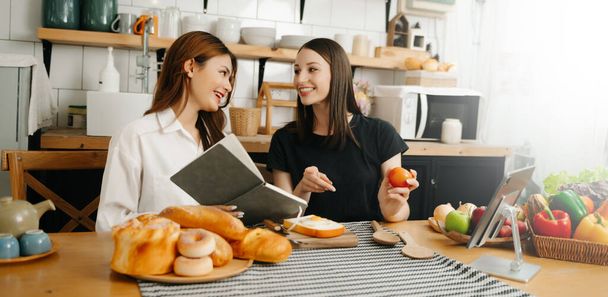 Two beautiful women in the kitchen in an apron, fresh vegetables on the table, writes down her favourite recipes, comes up with ideas for dishes  - 写真・画像