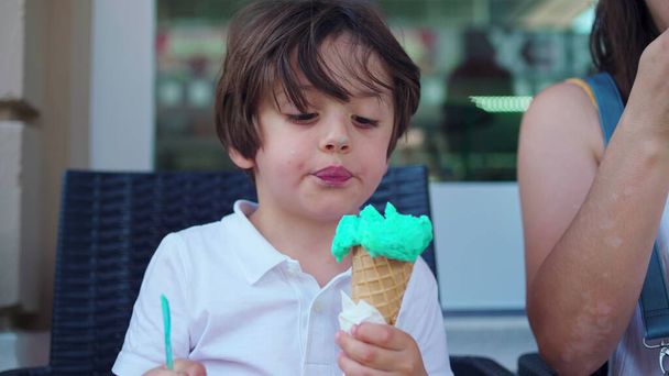 Kind genießt Eiszapfen im Parlo-Geschäft während des Sommertages. Mutter bringt Kegel näher an den Mund des kleinen Jungen, um zu verhindern, dass Kleidung beschmutzt wird - Foto, Bild