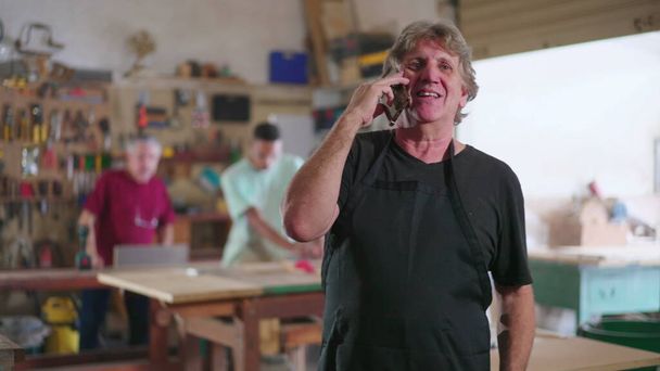 Μικρή επιχείρηση του εργαστηρίου ξυλουργικής μιλώντας με τον πελάτη στο κινητό ενώ αλληλεπιδρούν με τον εργαζόμενο στο παρασκήνιο που ασχολούνται με την ξυλουργική επιχείρηση - Φωτογραφία, εικόνα