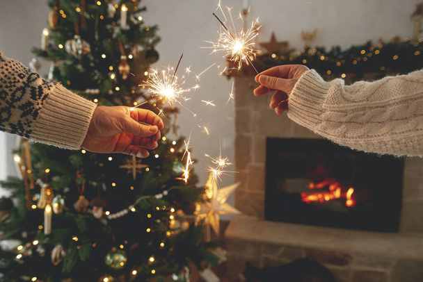 Szczęśliwego Nowego Roku! Płonące iskry w rękach na tle nowoczesnego wiejskiego kominka i choinki ze złotymi światłami. Fajerwerki świecące w dłoniach, para świętująca w świątecznym pokoju urządzone - Zdjęcie, obraz