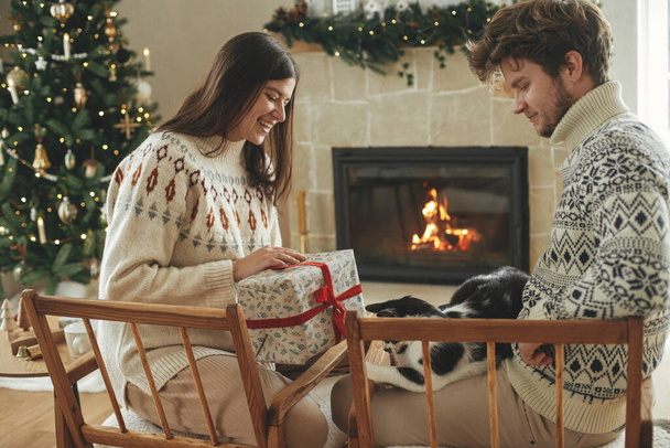 Szczęśliwa para w przytulnych swetrach bawiących się z kotem i wymieniających stylowe prezenty świąteczne na tle kominka z uroczystym płaszczem i nowoczesnej choinki ze światłami. Wesołych Świąt! - Zdjęcie, obraz