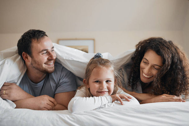 Love, happy and child in bed with her parents for bonding, relaxing and resting together with blanket. Smile, excited and portrait of girl kid laying with mother and father in bedroom of family home - Photo, Image