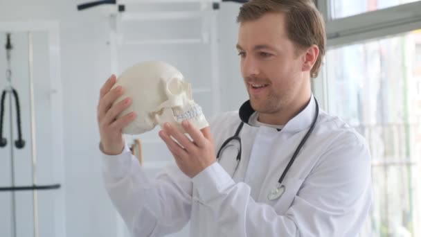 Increíblemente alegre joven y atractivo doctor sosteniendo un esqueleto de cráneo humano en sus manos. Un médico feliz y positivo en una clínica ortopédica moderna - Imágenes, Vídeo