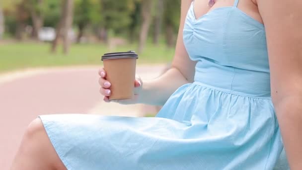 Dziewczyna w niebieskiej sukience pije kawę w parku. Kubek z kawą w rękach kobiety z bliska. Weź kawę na wynos. Napój pobudzający - Materiał filmowy, wideo