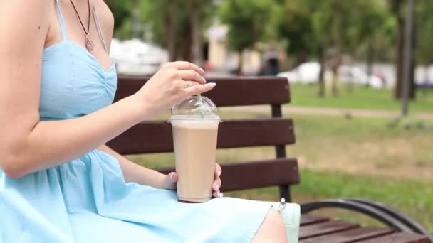 Een meisje in een blauwe jurk drinkt koffie in het park. Plastic beker met koffie in de handen van een vrouw close-up. Haal afhaalkoffie. Een verkwikkend drankje. IJslaag - Video