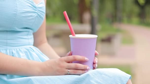 Uma rapariga de vestido azul bebe limonada no parque. Uma xícara de cor lavanda nas mãos de uma mulher close-up. Beba um copo para ir. Bebida refrescante - Filmagem, Vídeo
