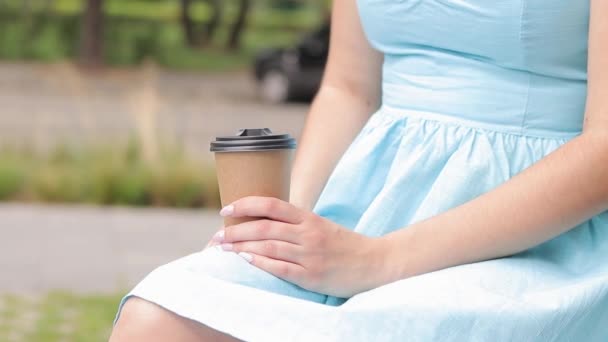 Een meisje in een blauwe jurk drinkt koffie in het park. Craft cup met koffie in de handen van een vrouw close-up. Haal afhaalkoffie. Versterkende drank - Video