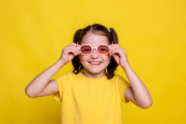 child in mockup yellow T-shirt in pink retro sunglasses and looking at camera on bright yellow background. studio advertising photoshoot - Photo, Image