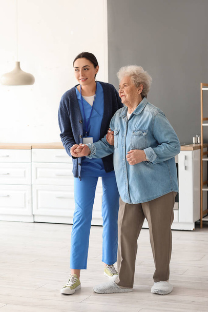 Jonge verzorger helpt oudere vrouw om te lopen in de keuken - Foto, afbeelding