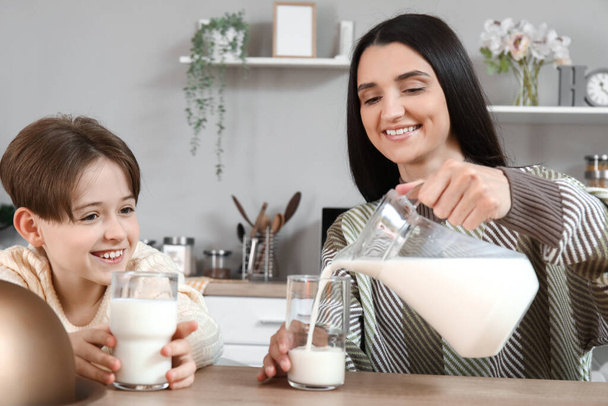 Happy little boy with his mother sitting at table and pouring fresh milk into glasses in kitchen - Photo, Image