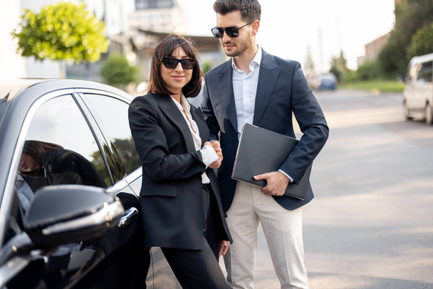 Biznesmen i bizneswoman ściskający sobie dłonie, stojący przed samochodem na zewnątrz. Koncepcja udanej transakcji i partnerstwa - Zdjęcie, obraz