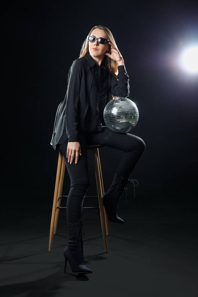 Beautiful young woman sitting on chair with disco ball against black background - Photo, Image