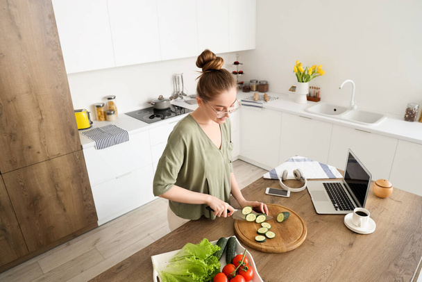Beautiful young woman cutting cucumber at table with modern laptop in light kitchen - Photo, Image