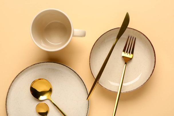 Чистая тарелка, миска, чашка и набор золотых столовых приборов на бежевом фоне - Фото, изображение