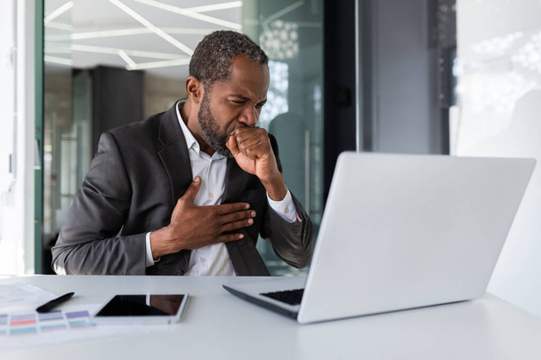 Enfermo adulto maduro afroamericano jefe toser en el lugar de trabajo dentro de la oficina, hombre de negocios que trabaja con el ordenador portátil sentado en el escritorio. - Foto, imagen