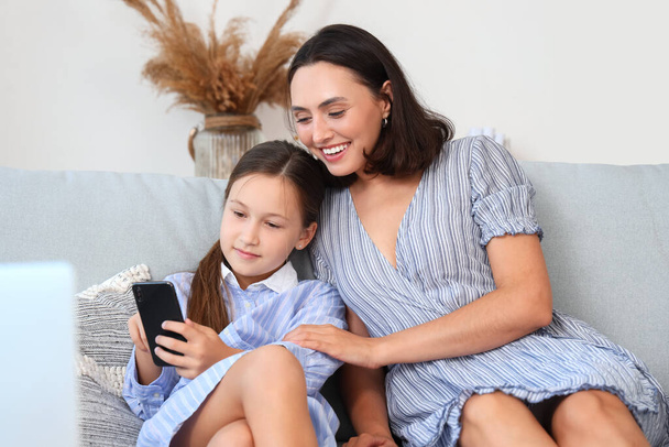 Madre e hija con teléfono móvil descansando en sofá gris en la sala de estar - Foto, Imagen