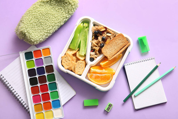 Lunchbox z pysznym jedzeniem i różnymi artykułami piśmiennymi na liliowym tle - Zdjęcie, obraz
