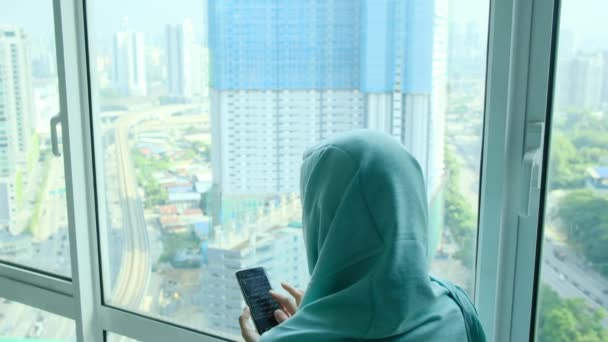 窓の近くの自宅でスマートフォンを使用してヘッドスカーフヒジャブの若いアラブイスラム教徒の女性 - 映像、動画