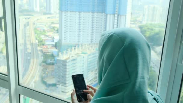 joven árabe musulmana mujer en hijab pañuelo para la cabeza usando el teléfono inteligente en casa cerca de la ventana - Metraje, vídeo