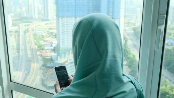 窓の近くの自宅でスマートフォンを使用してヘッドスカーフヒジャブの若いアラブイスラム教徒の女性 - 映像、動画