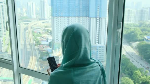 молодая арабская мусульманка в хиджабе в платке с помощью смартфона у окна - Кадры, видео