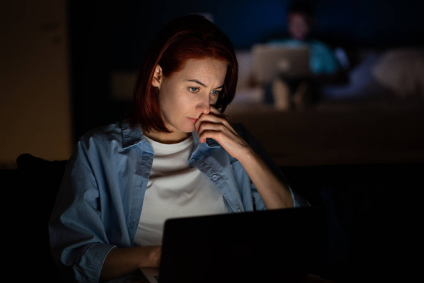 Poważna kobieta pracoholiczka pracująca na laptopie w ciemnościach w domu, patrząca na ekran. Praca online na komputerze. Wolny strzelec, odległa praca. Kobieta z uwagą tworząc pomysły myśląc dokumenty do czytania. - Zdjęcie, obraz