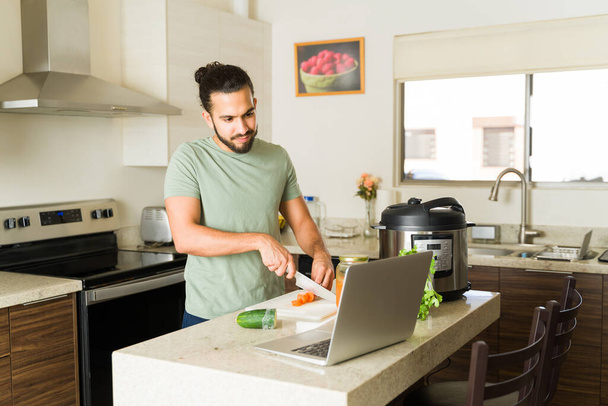 Handsome mexican man cutting vegetables in the kitchen and cooking food for lunch while watching a video tutorial on the laptop following a recipe  - Photo, Image