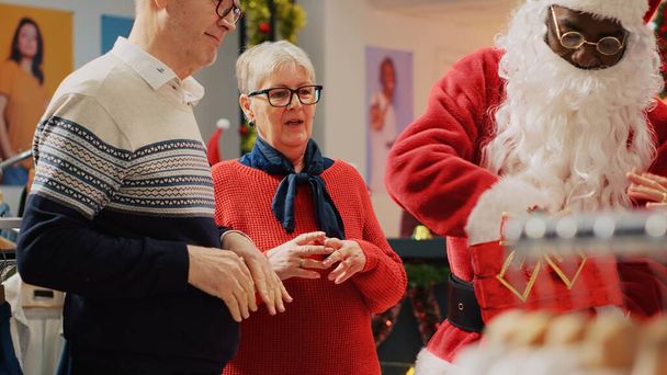 Klienci uczestniczący w loterii świątecznej posiadają przez sprzedawcę przebranego za Świętego Mikołaja w świątecznym sklepie odzieżowym. Szczęśliwa para starców podekscytowana po wygraniu konkursu promocyjnego sklepu z modą - Zdjęcie, obraz